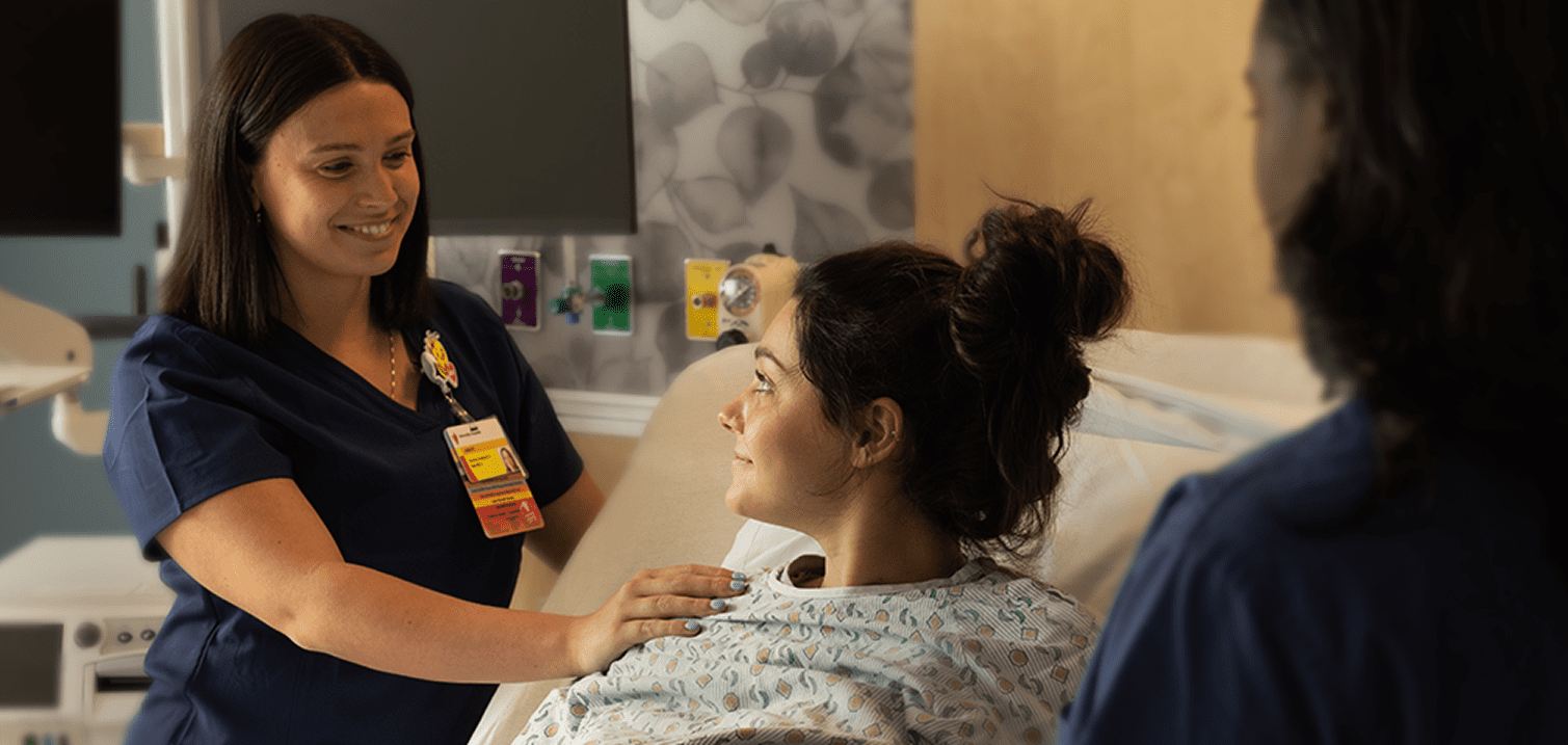 A smiling nurse in navy scrubs comforting a patient in a hospital bed while another healthcare professional looks on.
