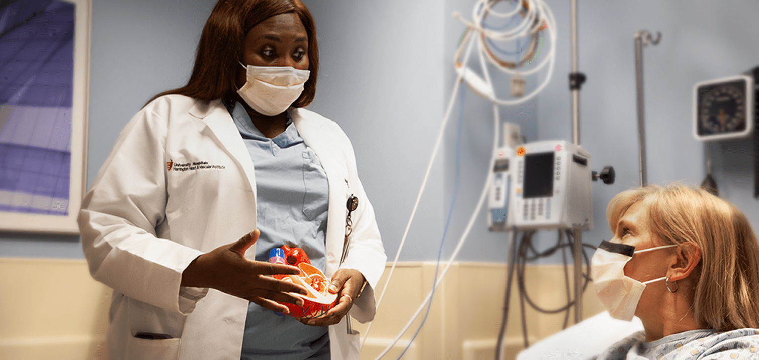 A masked healthcare professional in a white coat uses a heart model to explain a procedure to a patient in a hospital room.