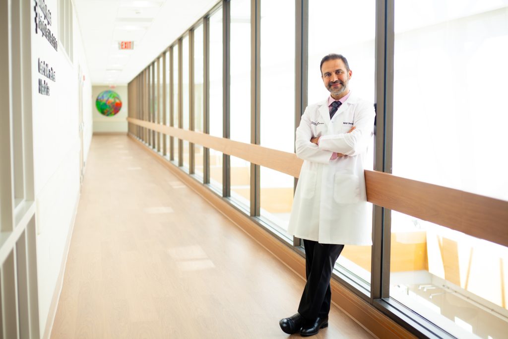 24060fde2ea985a8b433208a6c8ff407f0676699 A confident doctor in a white coat stands in a well-lit hospital corridor with a welcoming smile
