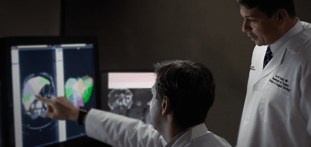 856bc365c7c72dcfc472fd0dcdc98e4042fe048e Two doctors in white coats analyze and discuss medical imaging scans on a computer screen in a dimly lit room.