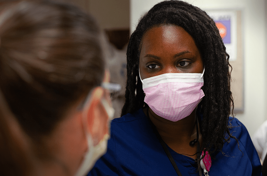 83394544c19770651c0e97fc7a06d45328f95742 A healthcare professional in navy scrubs wearing a pink face mask attentively talking with a colleague in a clinical setting.