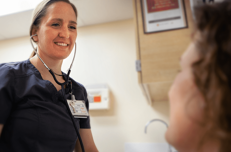 34035204ff34d56a45eb433a4ac1b241157acd25 A smiling nurse in navy scrubs uses a stethoscope to check on a patient in a medical examination room.