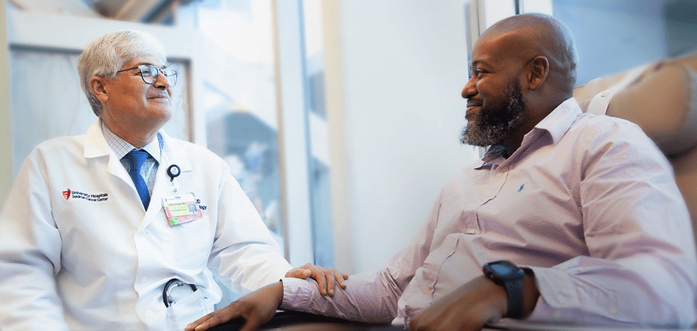 A doctor in a white coat warmly holding a patient's hand and engaging in conversation in a bright clinical setting.