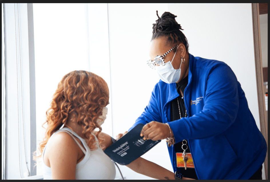 healthcare worker in blue uniform and mask fitting a blood pressure cuff on a seated patient’s upper arm near a window